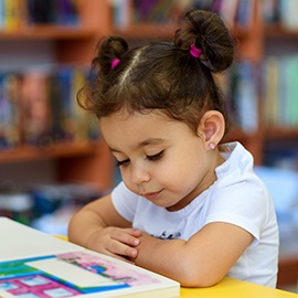 Little Girl Indoors In Front Of Books. Cute Young Toddler Sitting On A Chair Near Table and Reading Book. Child reads in a bookstore, surrounded by colorful books. Library, Shop, Shelving In Home.