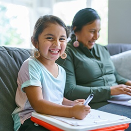 Portrait of a cute elementary age Native American girl laughing and looking directly at the camera while sitting on the couch and coloring. The little girl's mom is sitting next to her and is using a laptop computer.