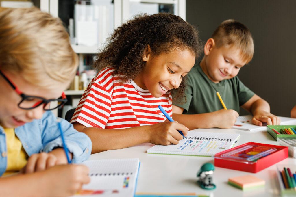 Positive ethnic student girl smiling and writing down data into notebook while sitting at table near classmates during test at school