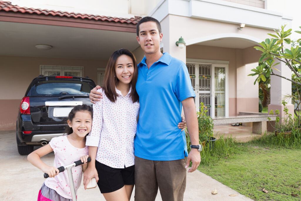 Young family of Thai and American descent poses for the camera in the front yard of their home in Nakhon Ratchasima, Thailand. The parents stand together with their 6 year old daughter and her scooter.