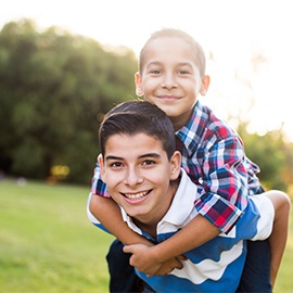 A teen boy carrying his younger brother on the back and smiling at the camera in a horizontal waist up shot outdoors.