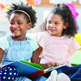 Three little girls discuss book together at preschool Three preschool age girls sit on the floor of their classroom together. They look at each other and smile as they discuss a book.