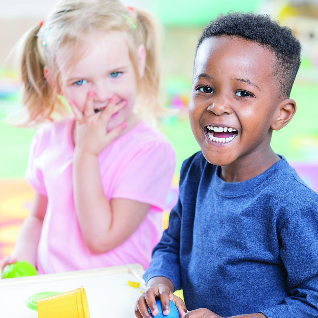 Adorable African American preschool boy smiles cheerfully as he plays with his friend in his preschool classroom.